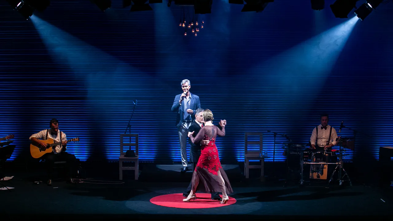 Espetáculo Tangos e Boleros no palco, com o cantor, um casal dançando tango e os músicos sob feixes de luz